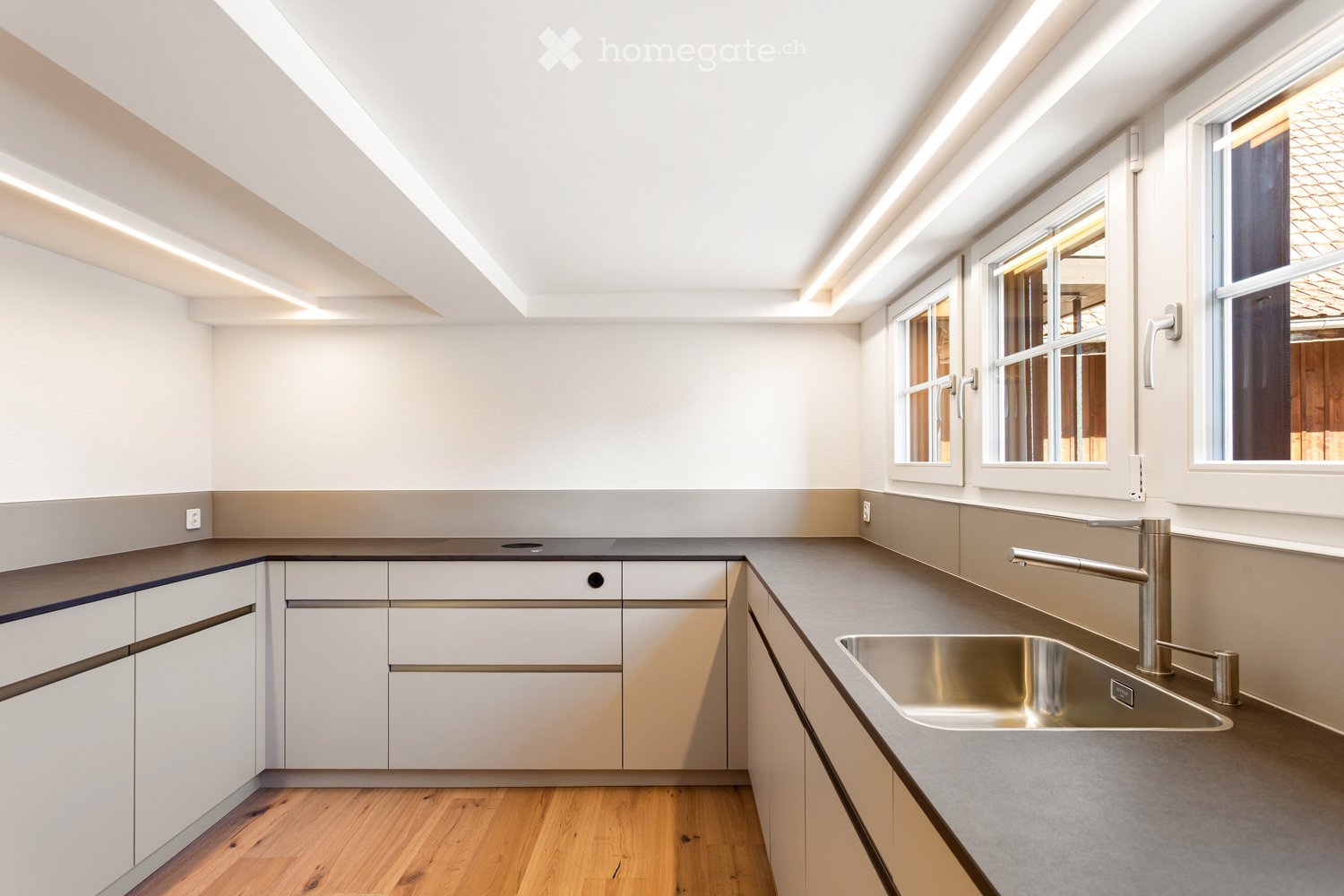 Modern kitchen with white cabinets, stainless steel sink, and sleek faucet. Natural light from large windows illuminates the space.
