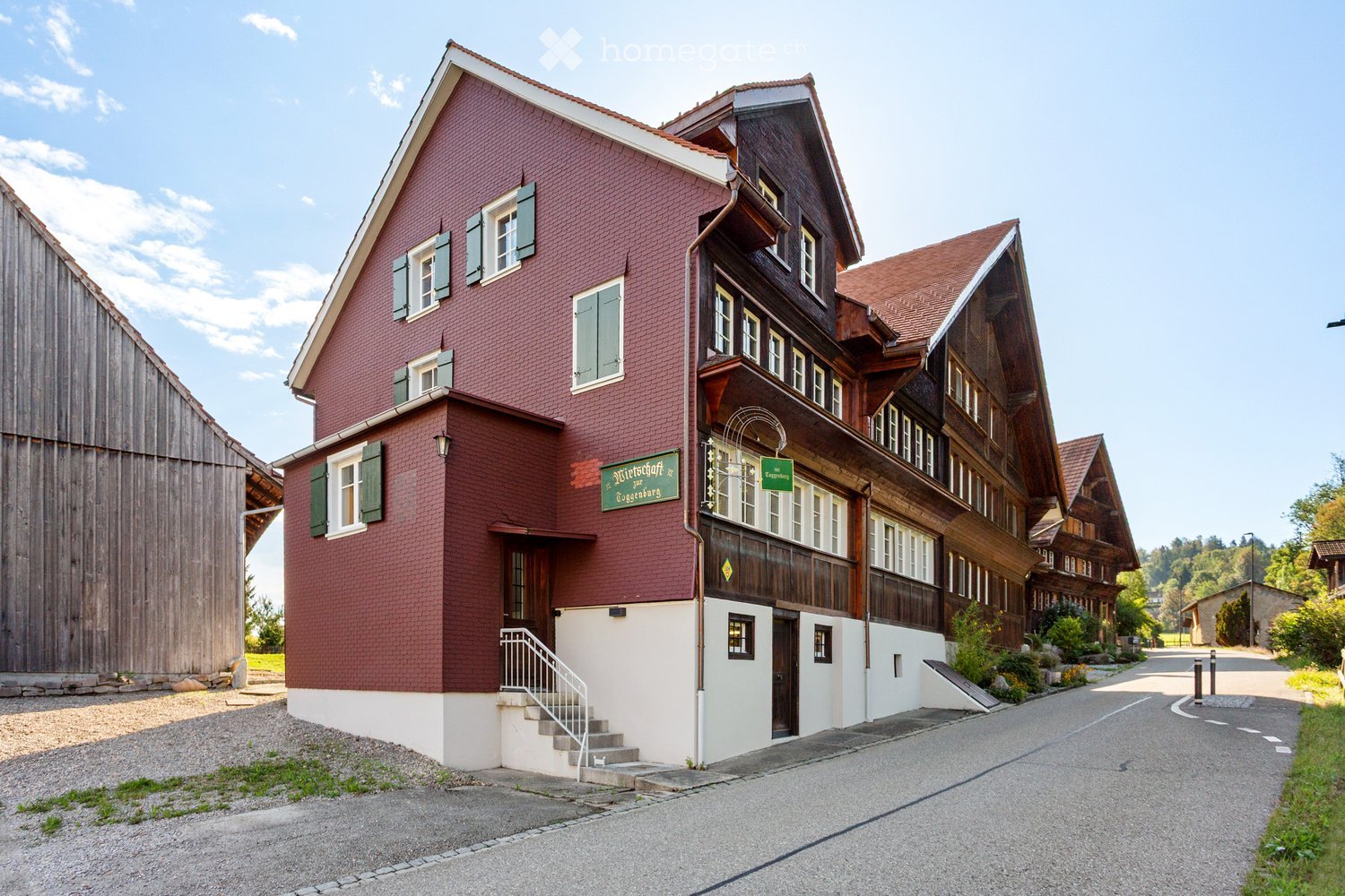Traditional two-story building with red brick facade and wooden elements, featuring multiple windows with shutters, a staircase leading to the entrance, and a signboard above the door.