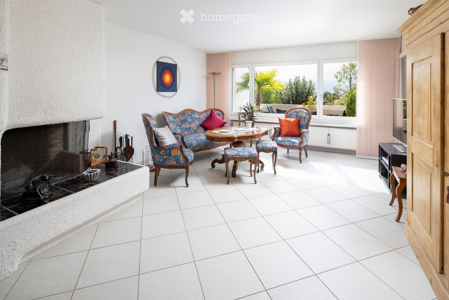 Bright living room with white tiled flooring, a fireplace on the left, a colorful sofa set, a small round table with chairs, and large windows offering a view of greenery outside.