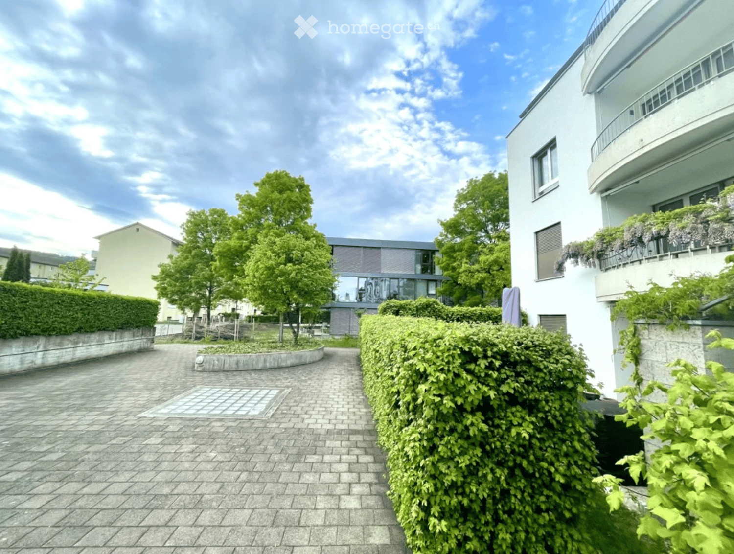 Garden area with a paved walkway, trimmed hedges, trees, and a modern white building with balconies and climbing plants.