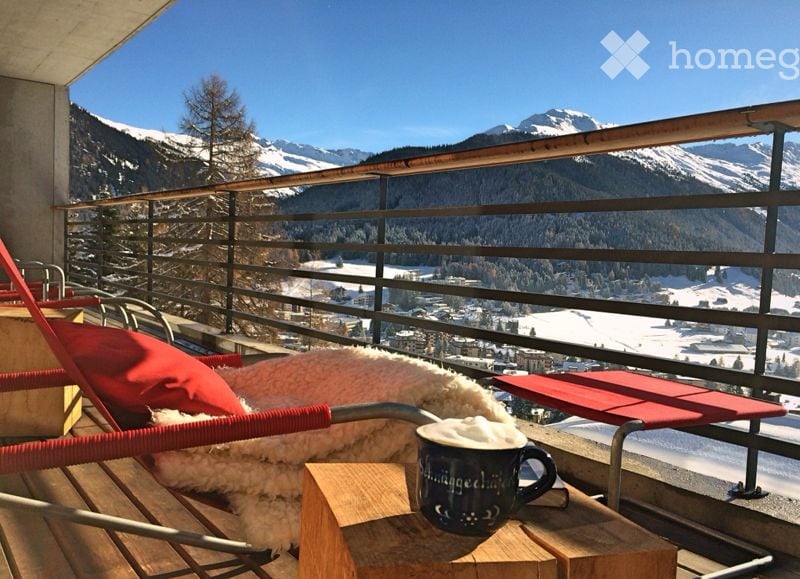 Balcony with a scenic mountain view, featuring a wooden table, a cup of hot chocolate, and a red lounge chair with a fur throw.