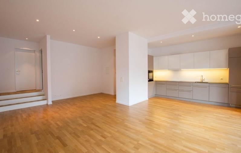 Bright, unfurnished room with wooden flooring, white walls, and a raised ground floor leading to a modern kitchen area with white cabinets and stainless steel appliances.