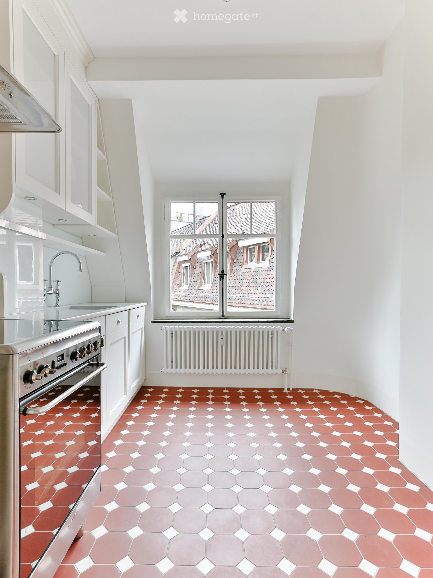 Modern kitchen with white cabinets, stainless steel oven, sink, and faucet. Features a large window with a view of rooftops and a radiator below. The floor has a geometric tile pattern in red and gray.
