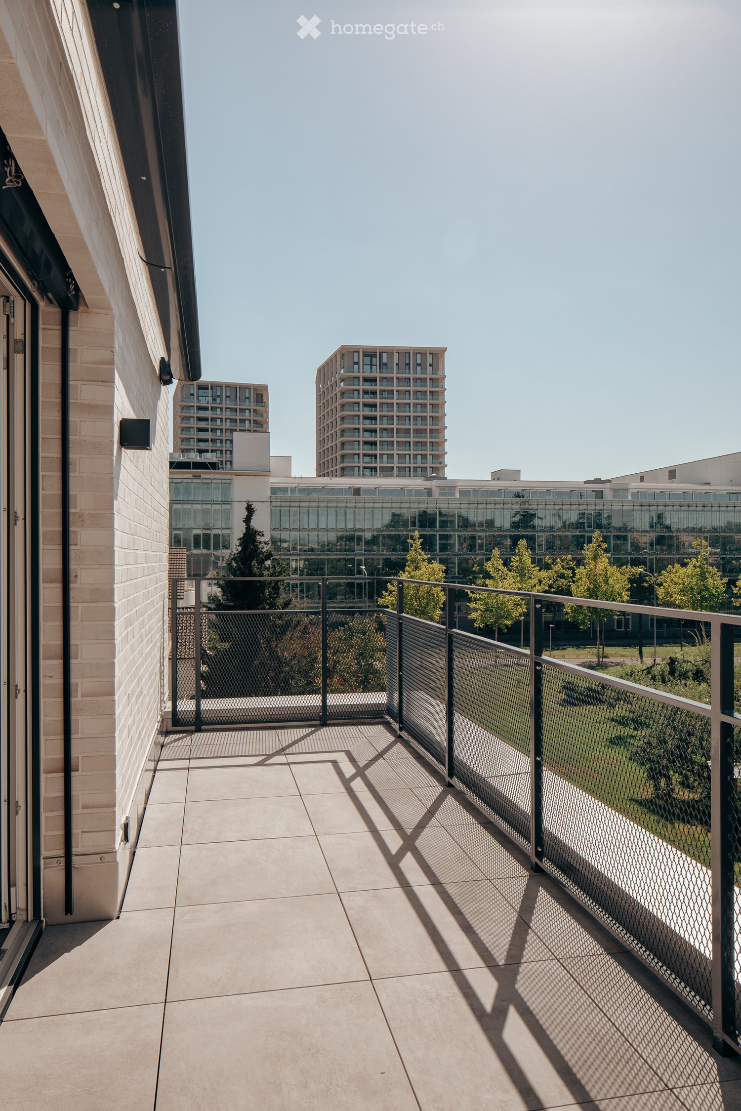 Balcony with tiled floor, metal railing, and view of modern buildings and greenery.