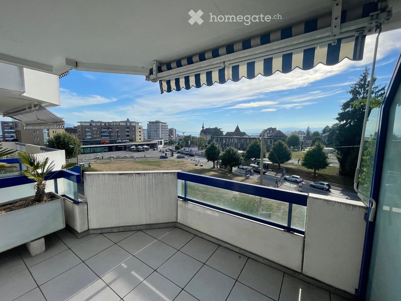 Balcony with tiled floor, glass railing, and potted plant, overlooking a cityscape with buildings, trees, and a parking area.