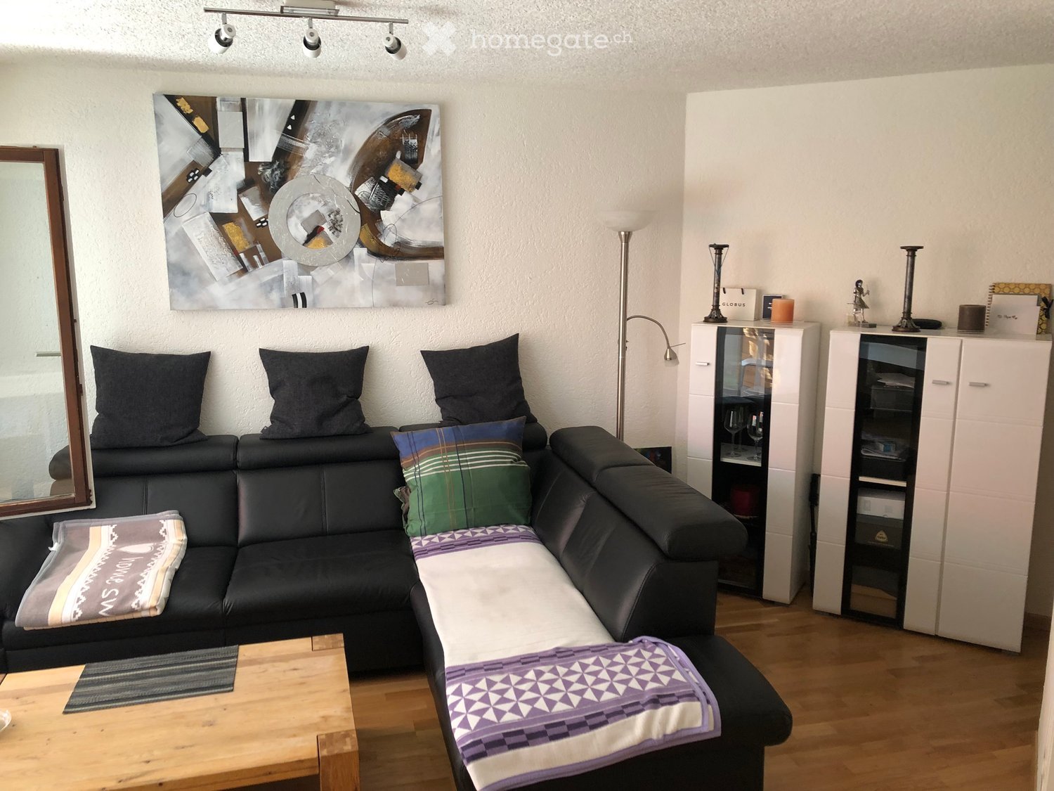 Modern living room with a black leather sofa, wooden coffee table, white cabinets, floor lamp, and artwork on the wall.
