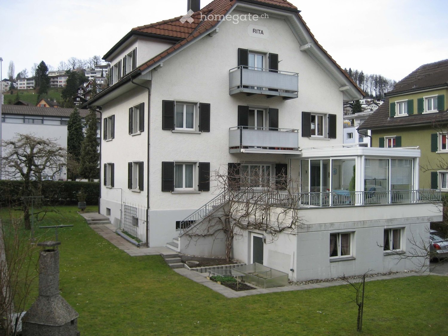 A two-story white house with a red-tiled roof, multiple windows with shutters, balconies, and a well-maintained garden with a stone chimney and steps leading to the entrance.