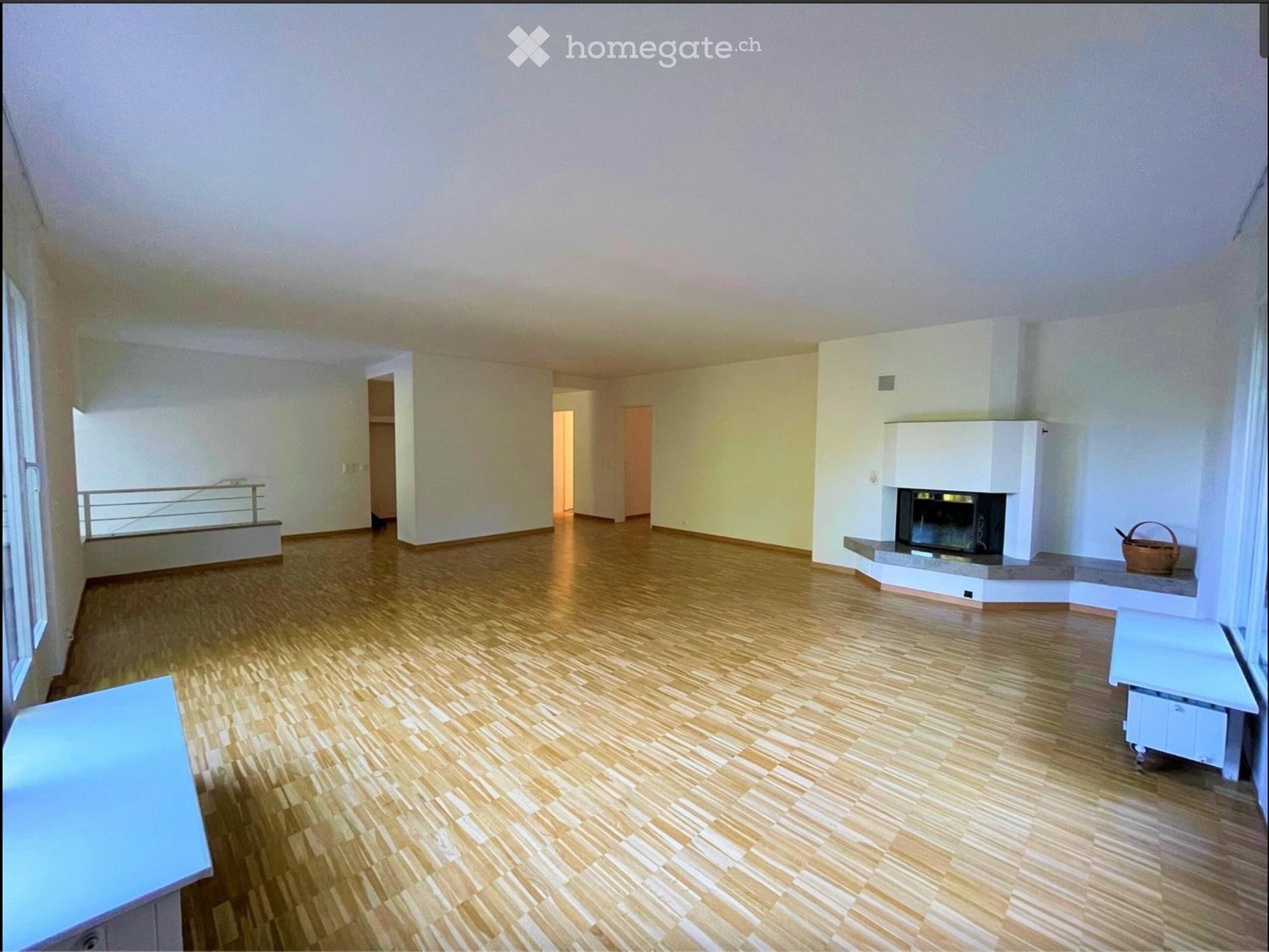 Empty living room with wooden flooring, white walls, fireplace, and large windows with a balcony view.