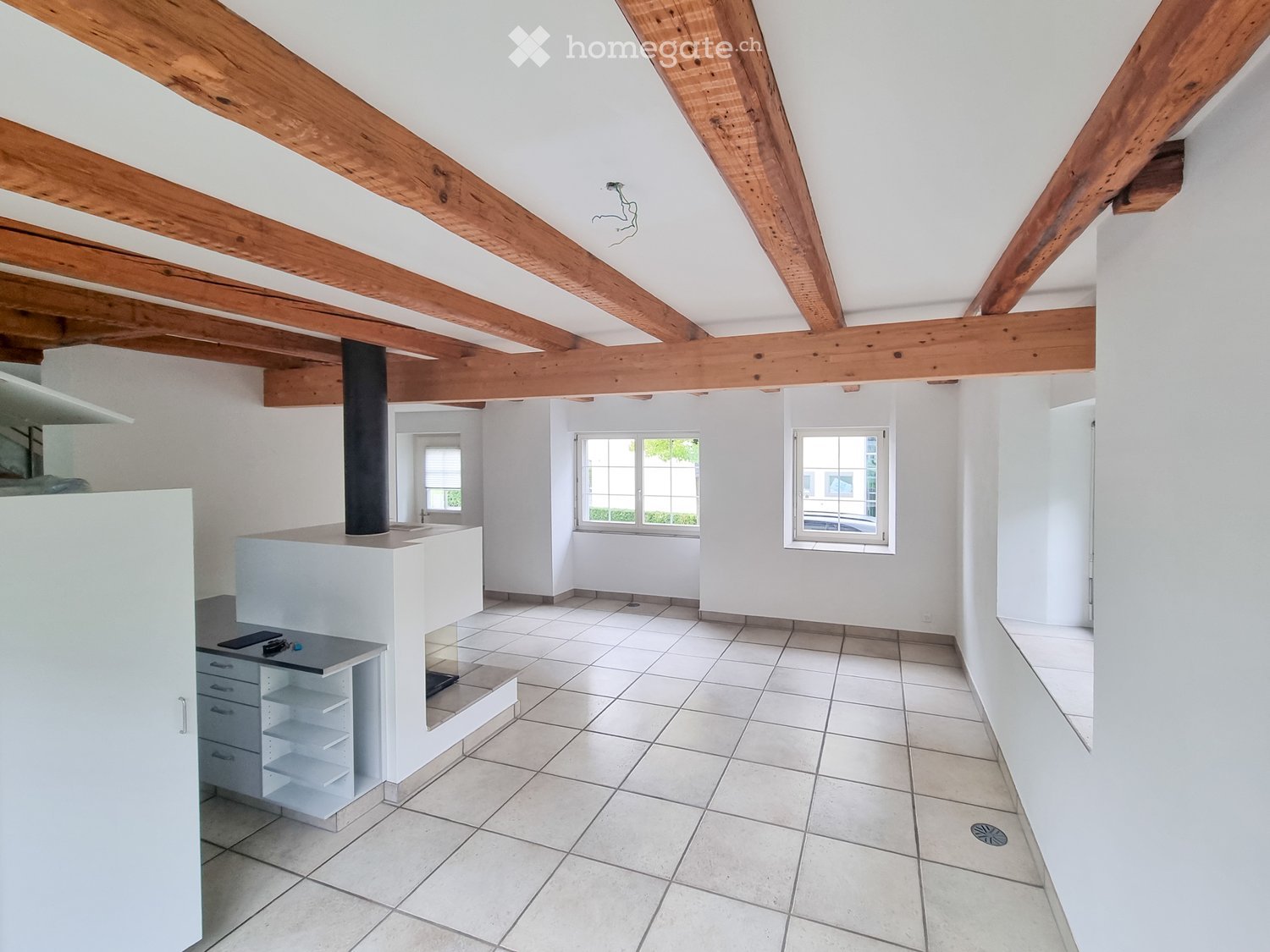 Bright, open room with exposed wooden beams on the ceiling, white tiled flooring, and large windows allowing natural light. Minimalist design with a small kitchen counter and built-in storage unit.