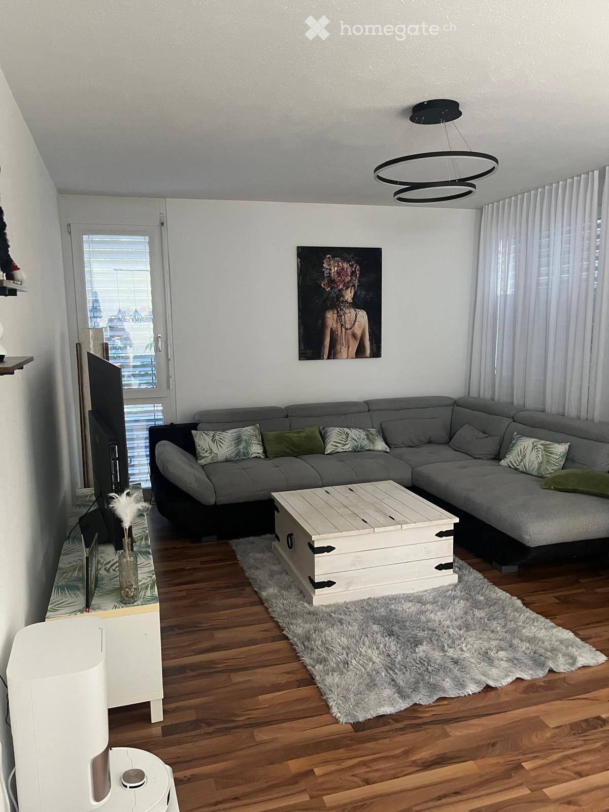 Modern living room with grey sectional sofa, wooden coffee table, fluffy rug, large painting, and TV stand.