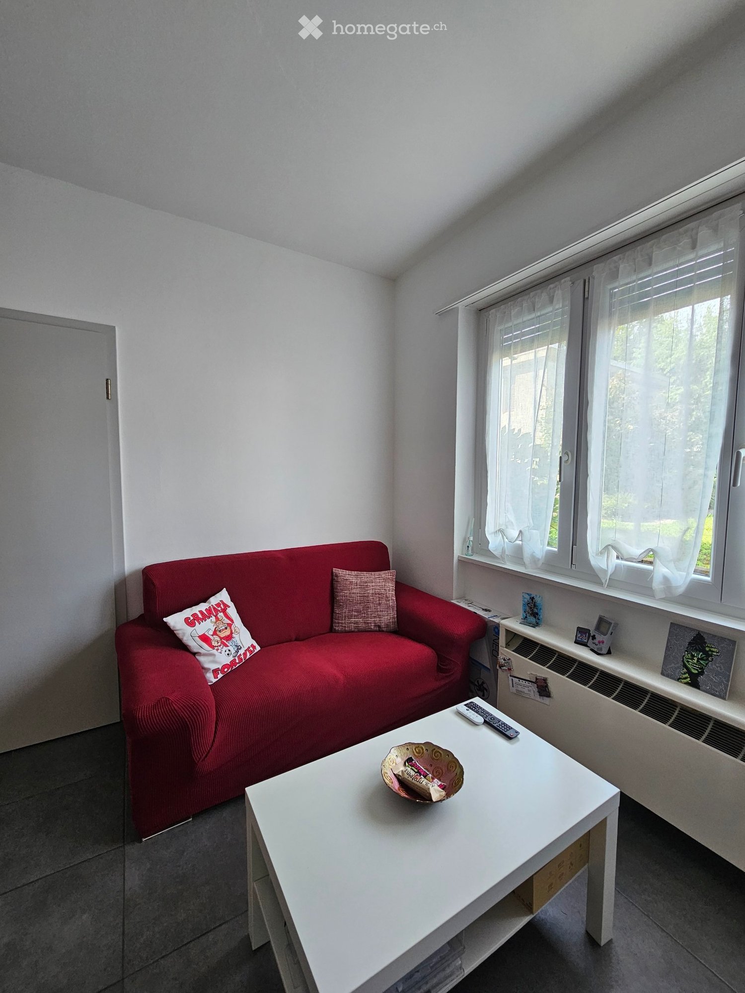 A modern living room with a red sofa, white coffee table, and large windows with white curtains. The room features a grey floor and white walls.