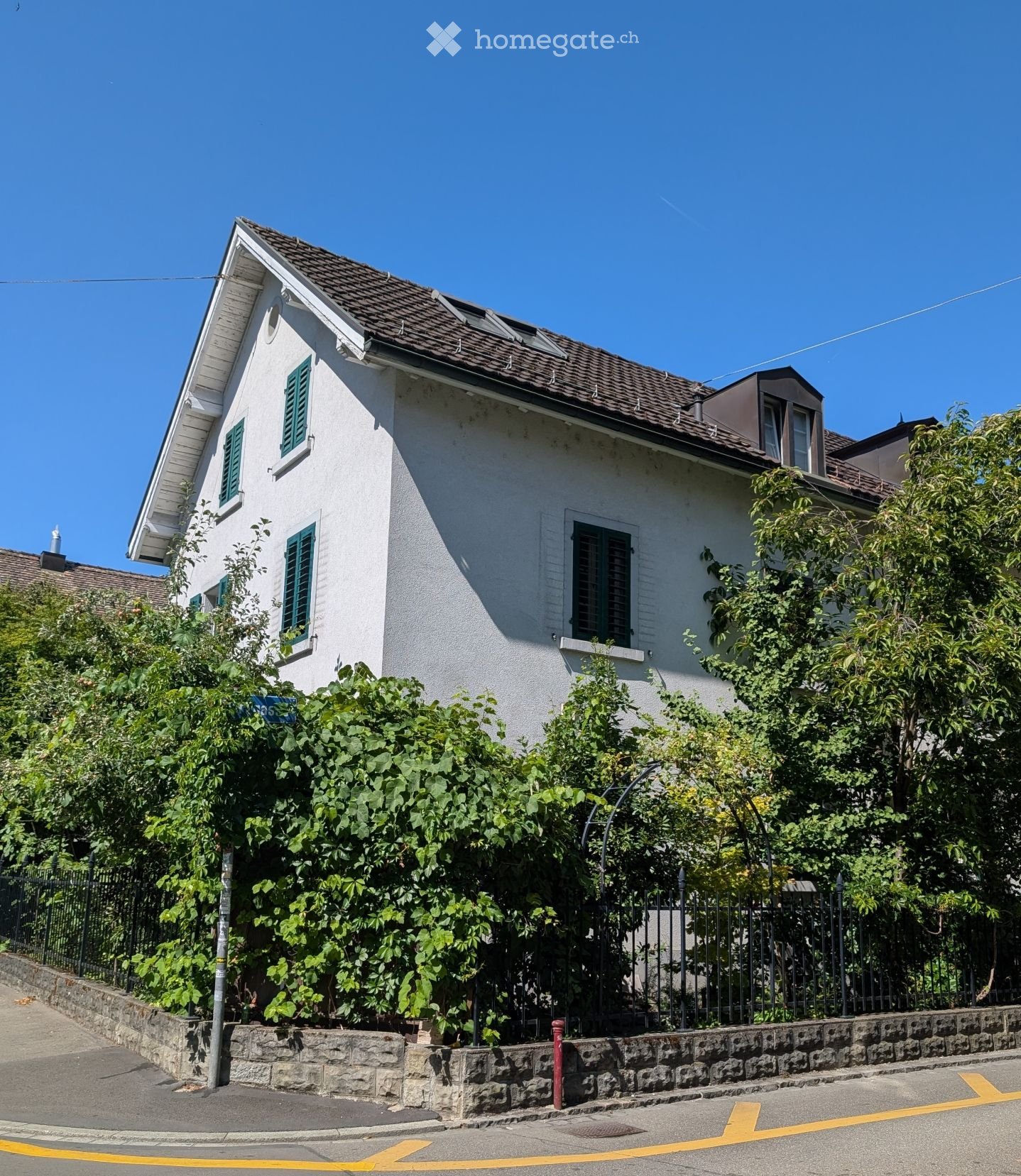 Two-story white house with green shutters, surrounded by lush greenery and a stone wall.