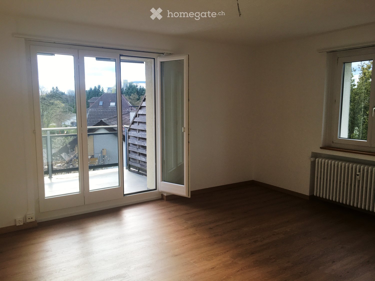 Empty room with wooden flooring, large sliding glass doors leading to a balcony, and a radiator on the wall.