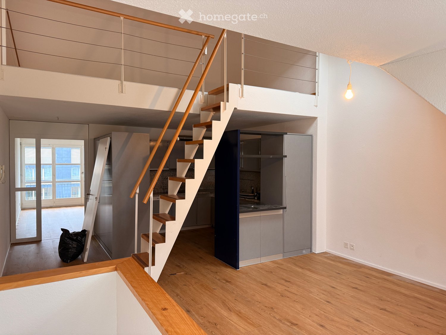 Modern hallway with wooden flooring, white walls, and a staircase leading to an upper level. Features include a sleek wooden railing, a small kitchen area with grey cabinets, and a large window with blinds.