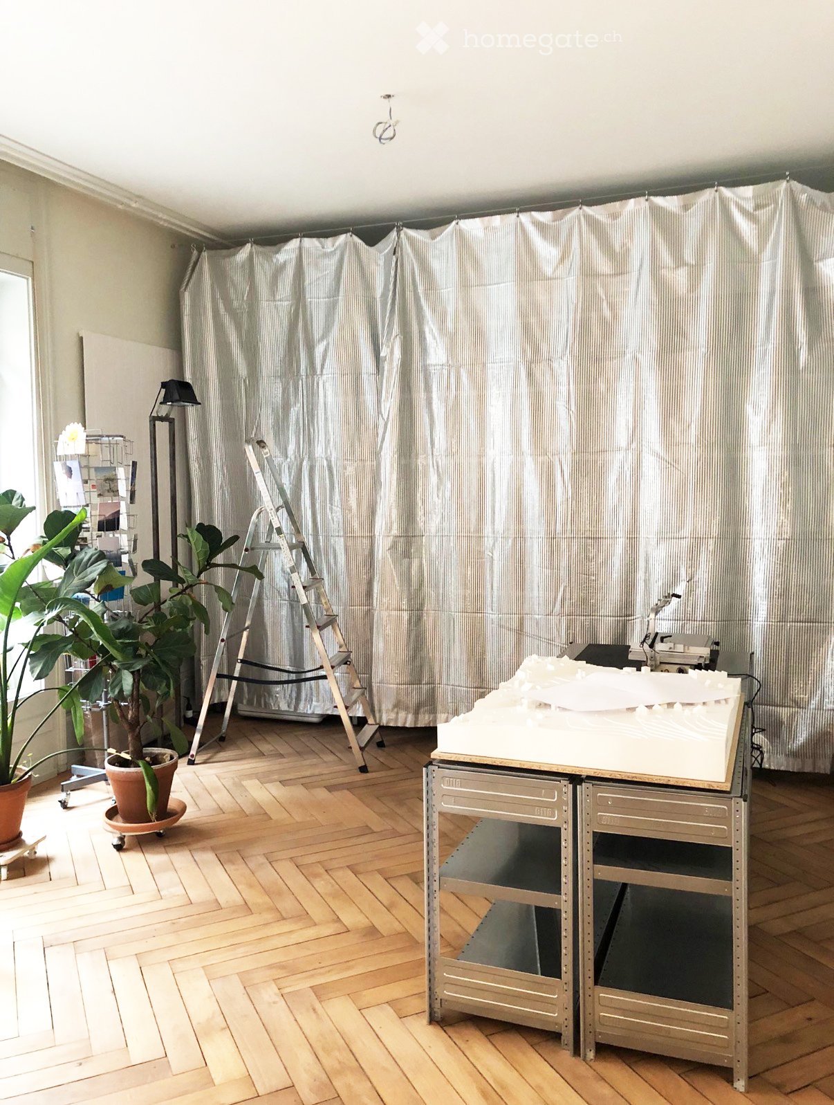 A room with wooden flooring, a large potted plant, a ladder, a table with a white object on it, and a lamp near the window.