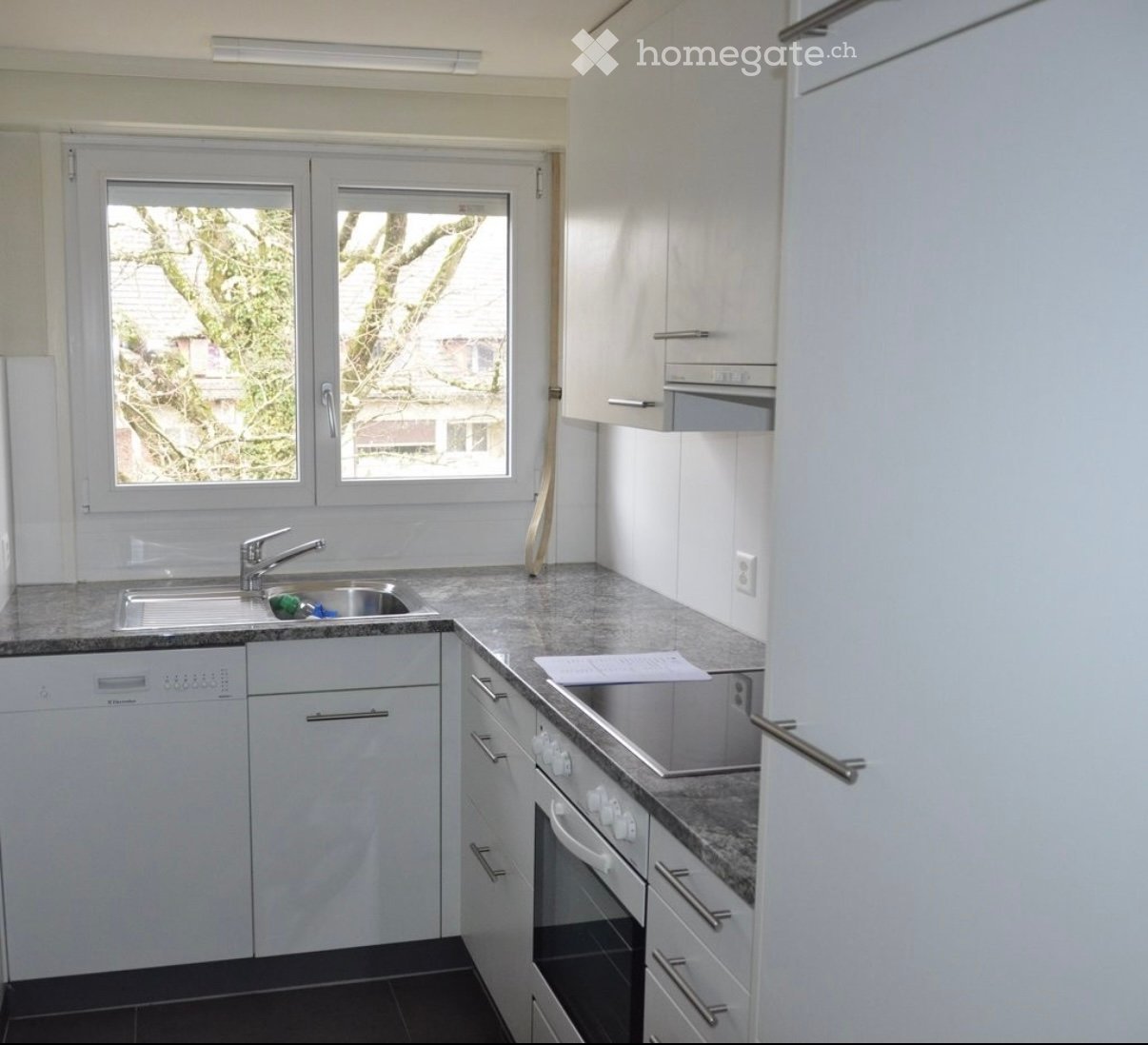 Modern kitchen with white cabinets, granite countertops, stainless steel sink, dishwasher, and oven. Large windows provide natural light.