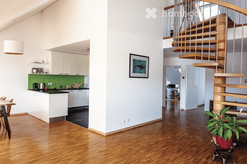 Open-plan living and kitchen area with light-colored cabinets, green backsplash, wooden flooring, a staircase with wooden steps and metal railings, and a dining table with chairs.