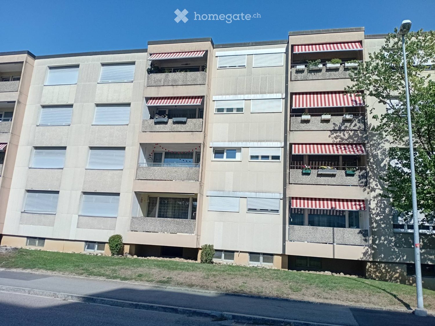 Multi-story residential building with beige facade, multiple windows, balconies with striped awnings, and small potted plants.