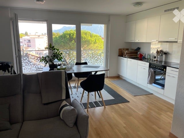 Open-plan living and dining area with a grey sofa, coffee table, dining table with chairs, and a modern kitchen with white cabinets, an oven, and a faucet. Large windows offer a scenic mountain view.