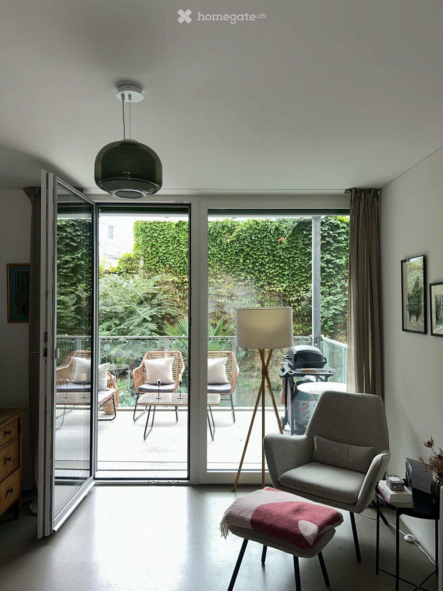 Bright living room with large glass doors opening to a balcony, featuring a grey armchair, a small table, a floor lamp, and a decorative stool with a pink throw.