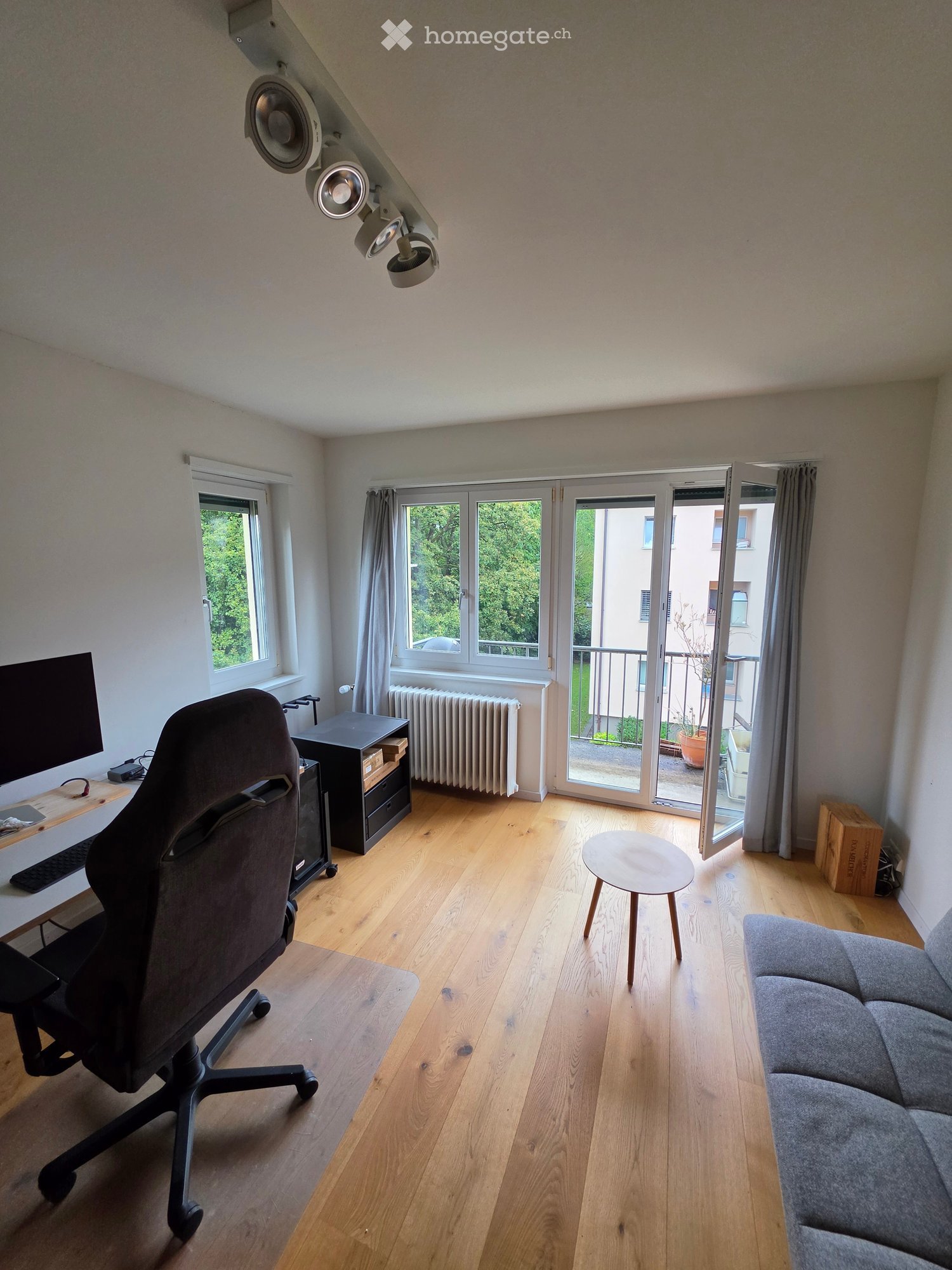 A modern, well-lit room with wooden flooring, large windows, and a balcony door. Features include a grey sofa, a round wooden stool, a desk with a screen and chair, and a radiator under the windows.