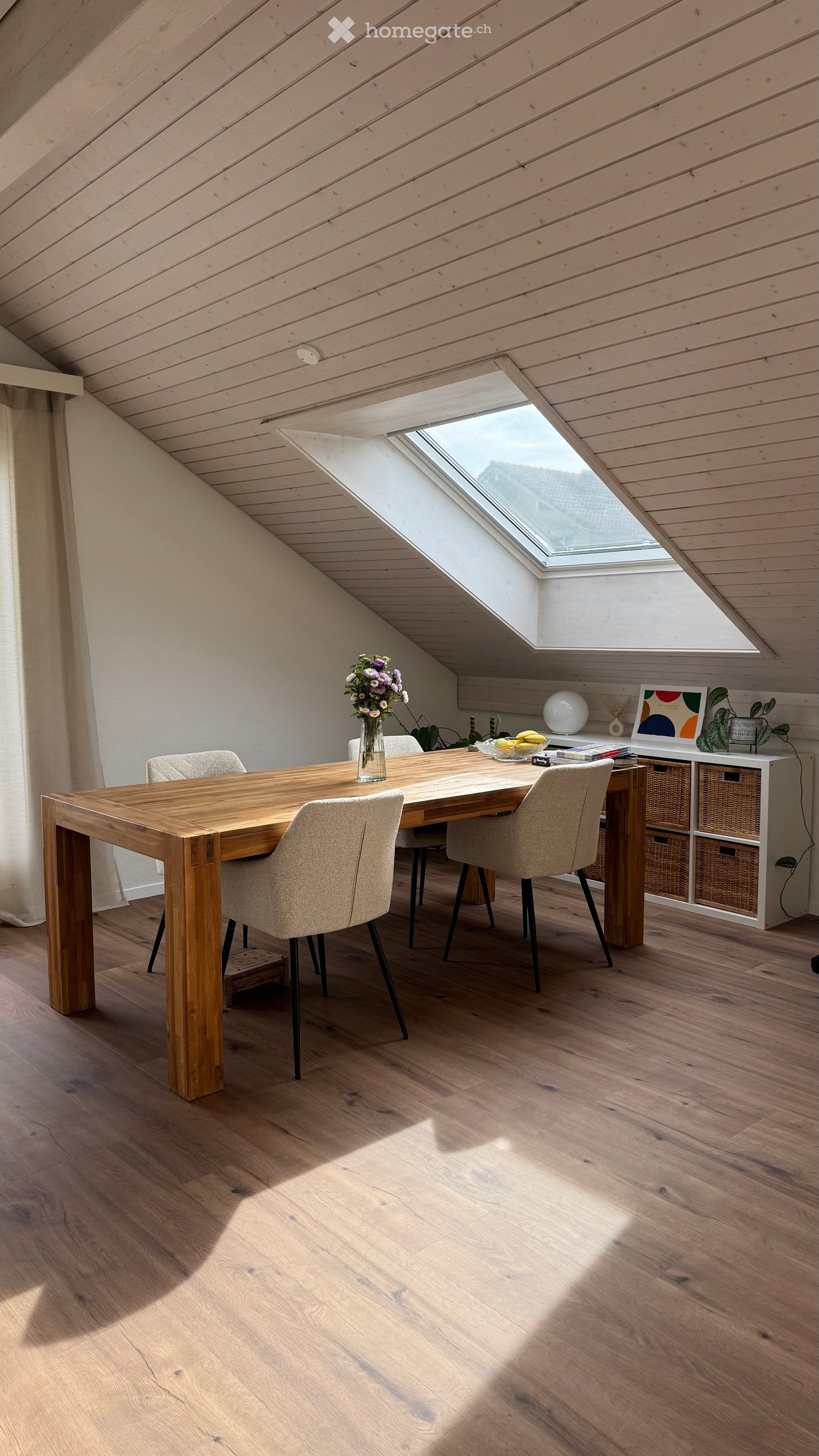 Bright dining area with wooden table, four chairs, vase with flowers, and a storage unit with books and decorative items.