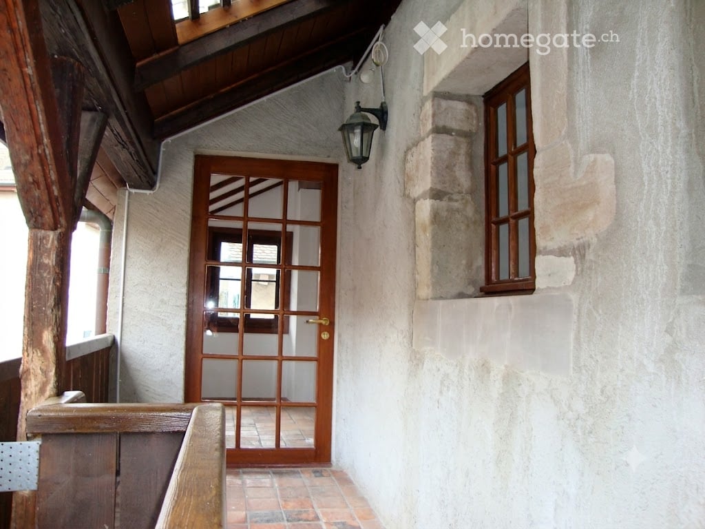 A narrow hallway with exposed wooden beams, a wooden door with glass panels, and a small window on the right side.