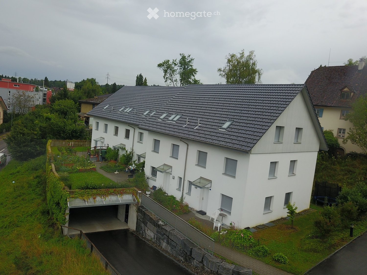 white house, gray roof, multiple windows, small garden, garage, several plants, paved driveway, small bridge
