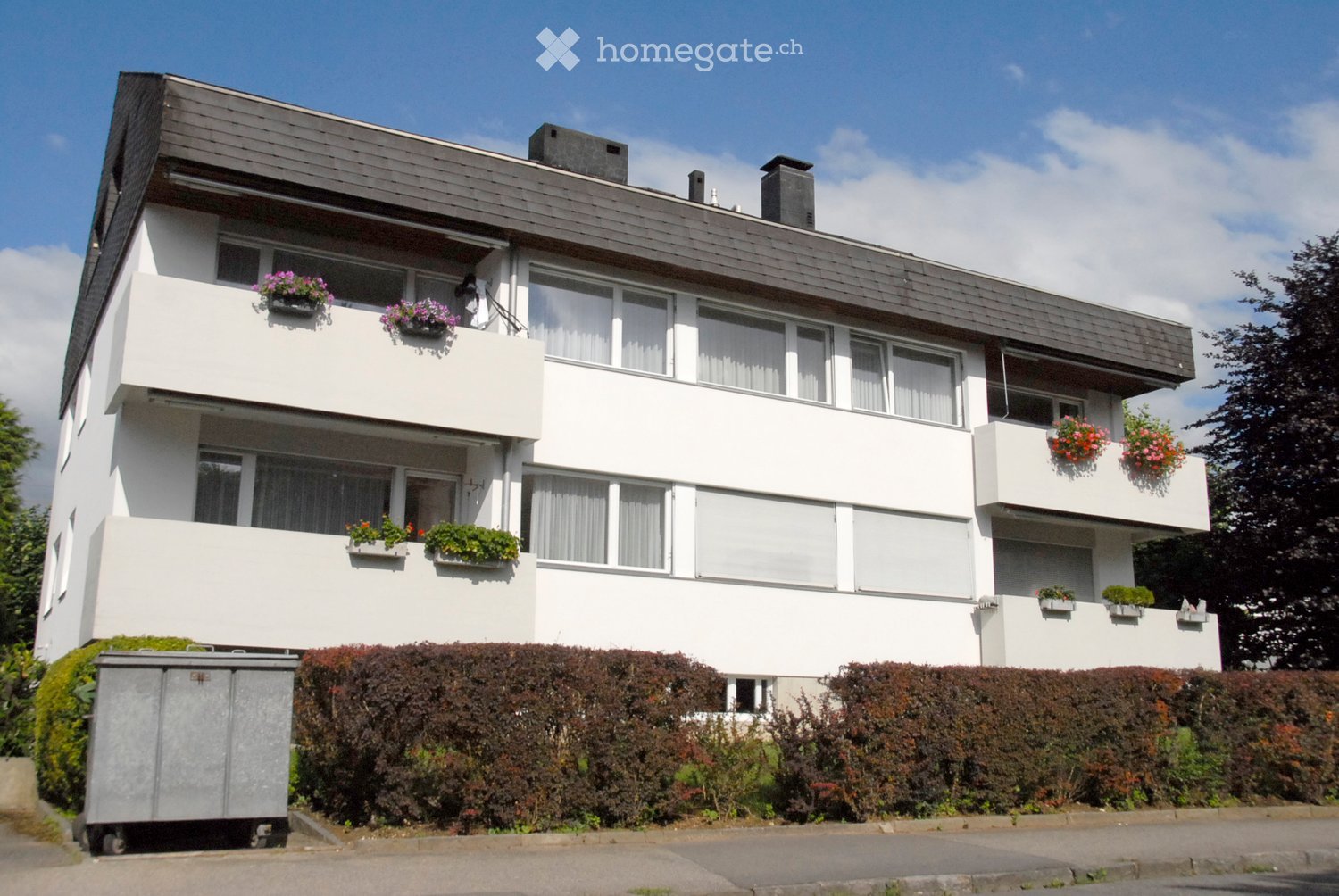 A modern, white multi-story house with balconies, large windows, and flower boxes, surrounded by greenery and a raised ground floor.