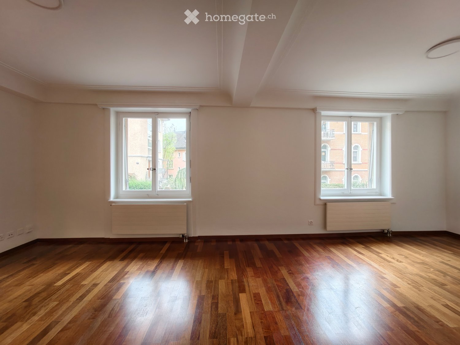 Empty room with wooden flooring, white walls, two windows with radiators, and a simple ceiling light fixture.
