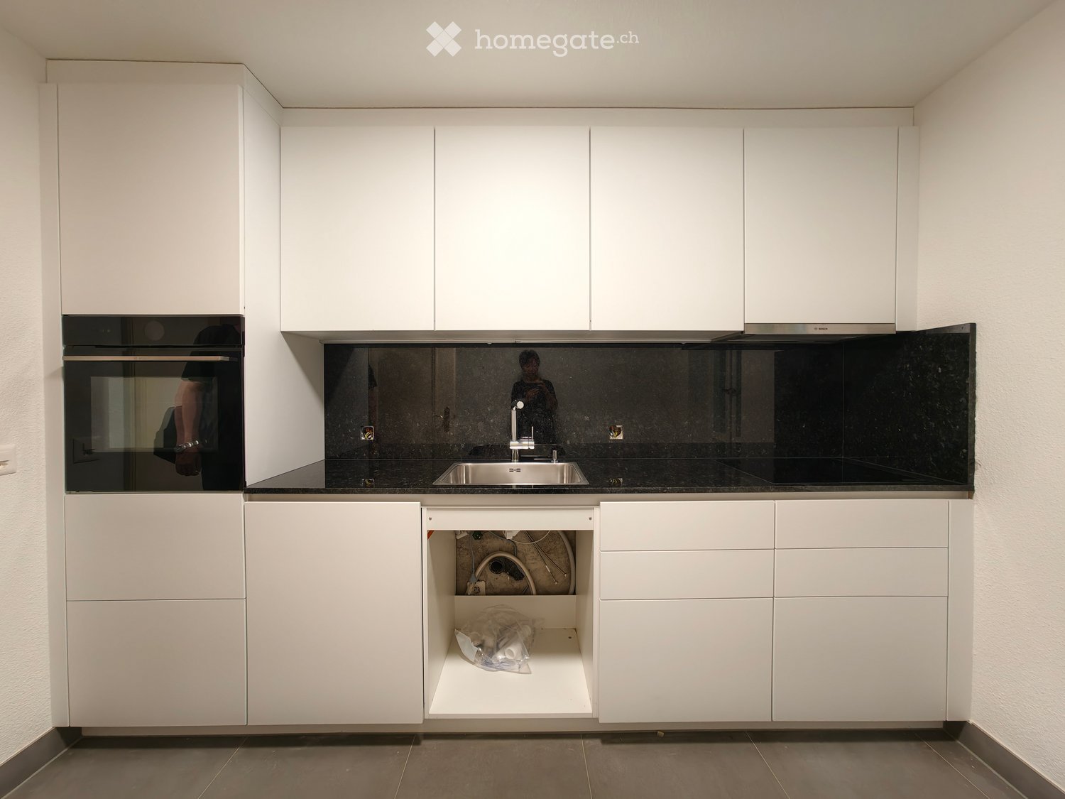 Modern kitchen with white cabinets, stainless steel sink, and built-in oven. Open storage compartment contains cleaning supplies.