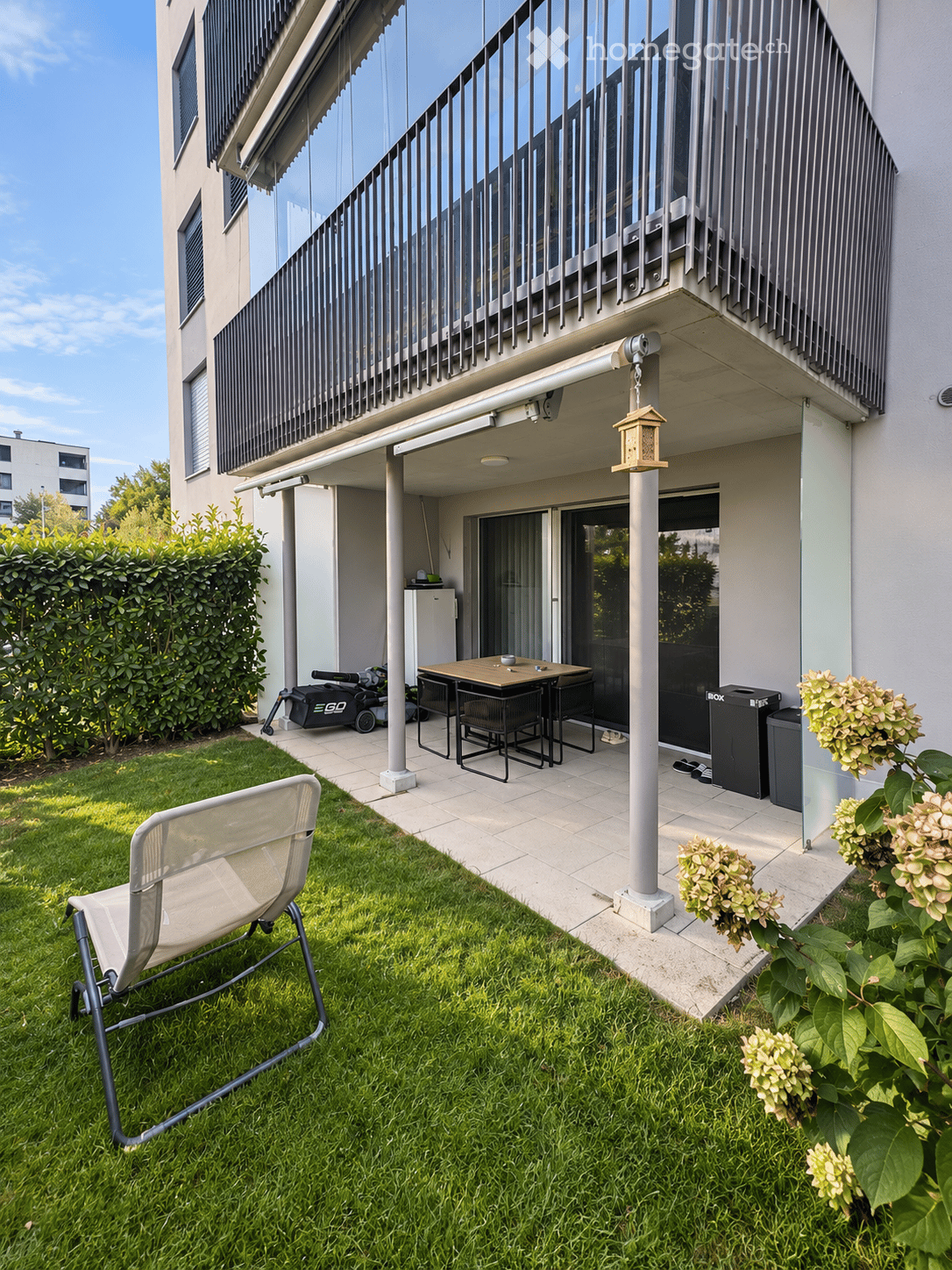 Garden area with a lounge chair, small table, and stools, surrounded by green grass and flowers, with a covered patio area and a balcony above.