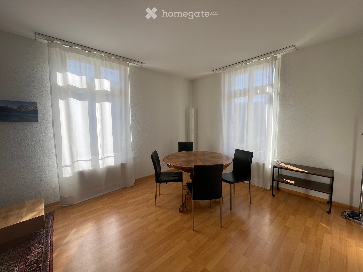 Dining room with wooden floor, large windows with white curtains, round wooden dining table with four black chairs, small shelf, and a painting on the wall.