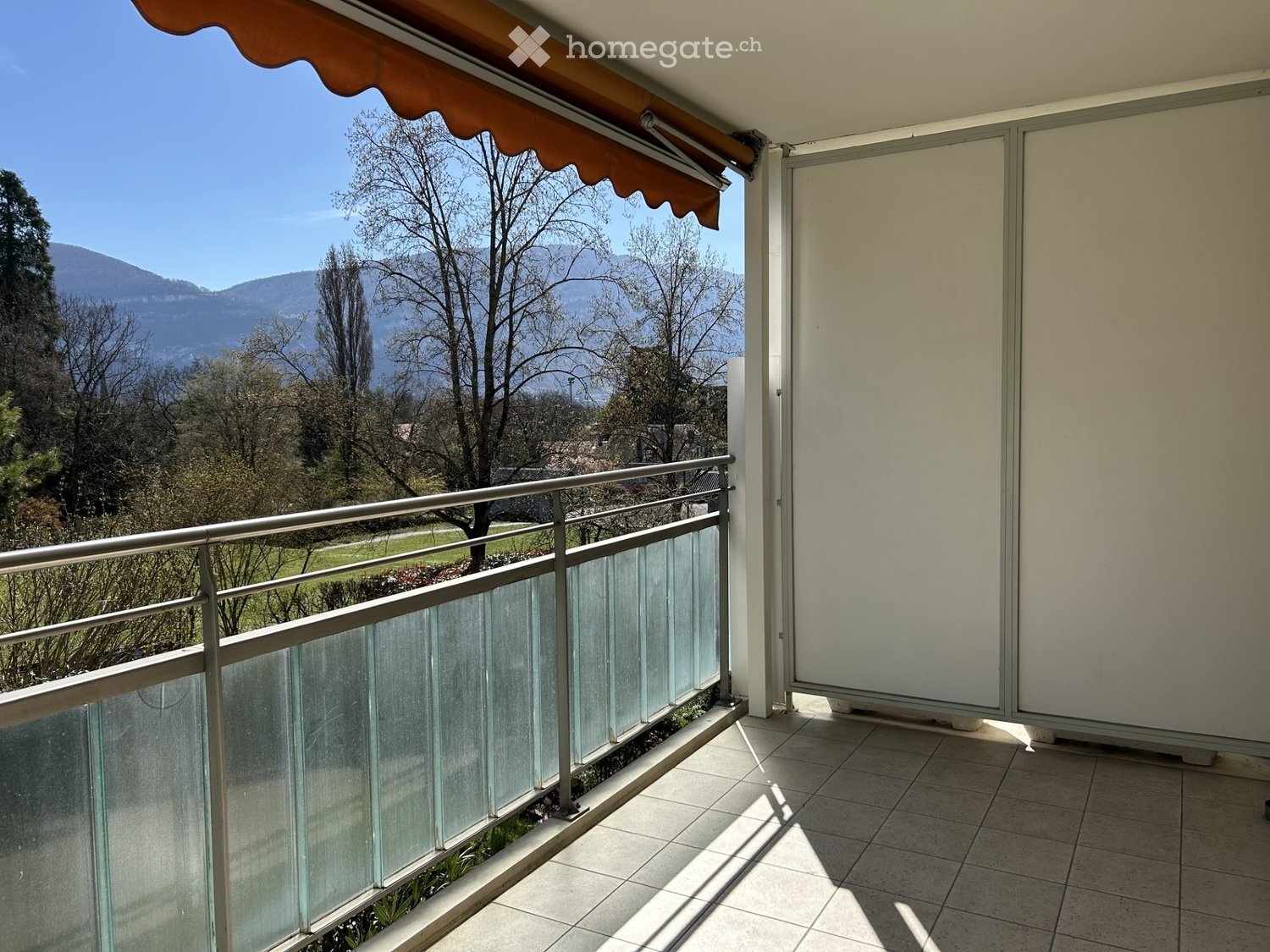 Balcony with tiled floor, glass railing, and retractable awning, overlooking a scenic landscape with trees and mountains.