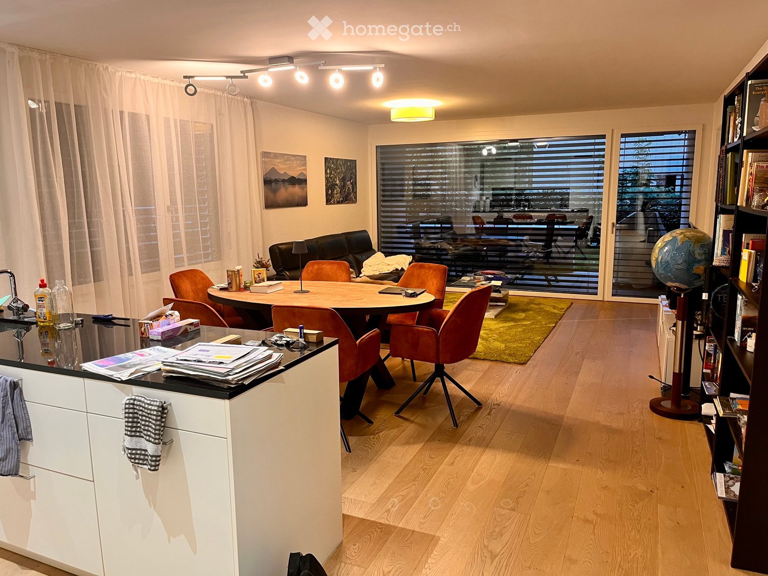 Modern living and dining area with a round wooden dining table, orange chairs, a black leather sofa, a bookshelf, and a globe. The space features a kitchen counter with a faucet, a curtain, and artwork on the walls.