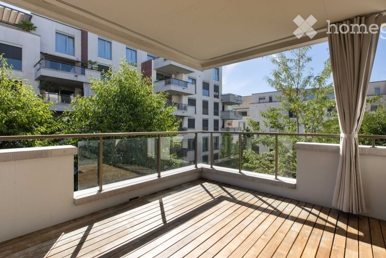 Spacious balcony with wooden decking, glass railing, and large windows offering a view of the city and surrounding greenery.