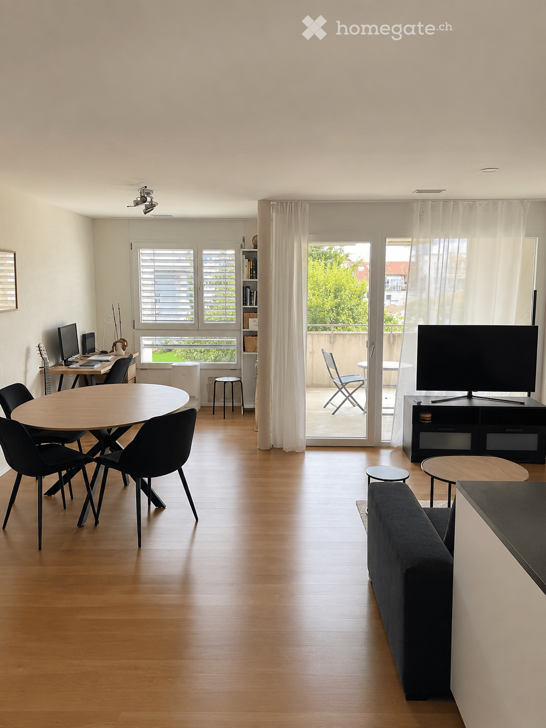 Modern living room with wooden flooring, large windows, and a balcony. Features a sofa, coffee table, dining table with chairs, and a TV stand.
