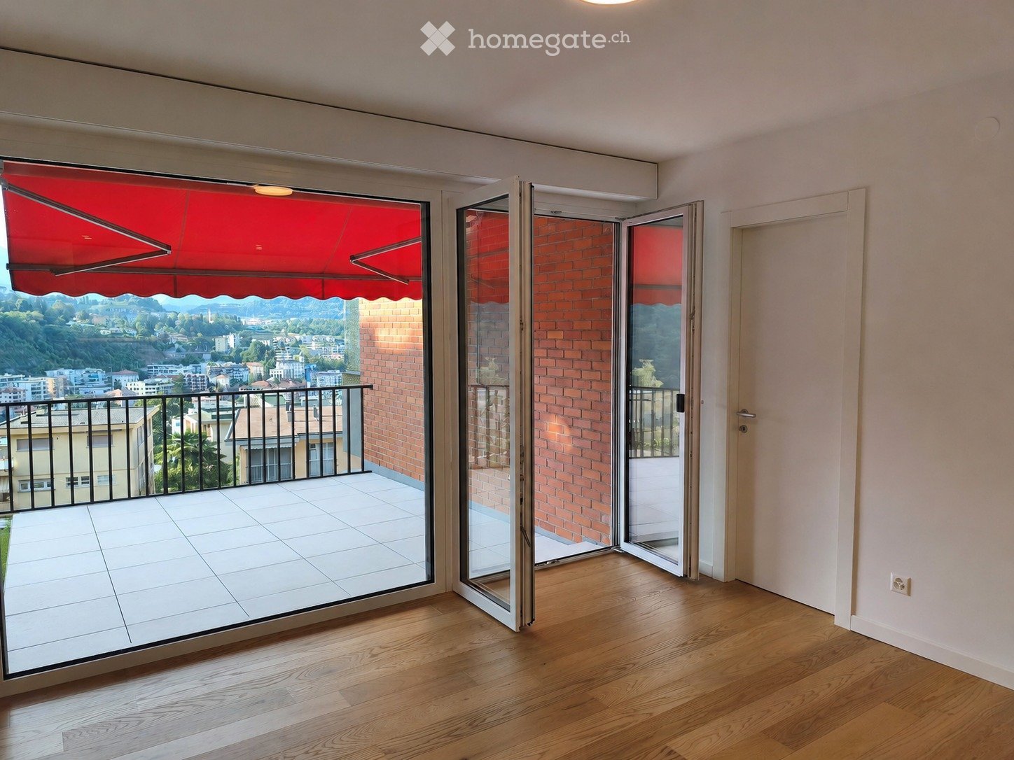 Spacious balcony with white tiled floor, black metal railing, and red awning. The balcony offers a scenic view of the city and surrounding hills.