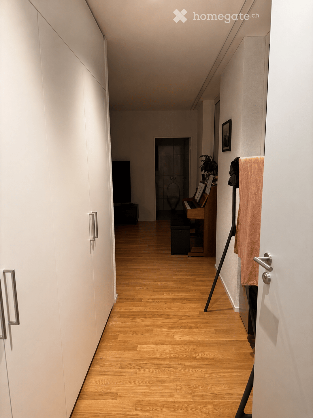 A narrow hallway with light wooden flooring, white walls, and built-in white cabinets on one side. A door leads to a bathroom, and a small plant and piano are visible in the adjacent room.