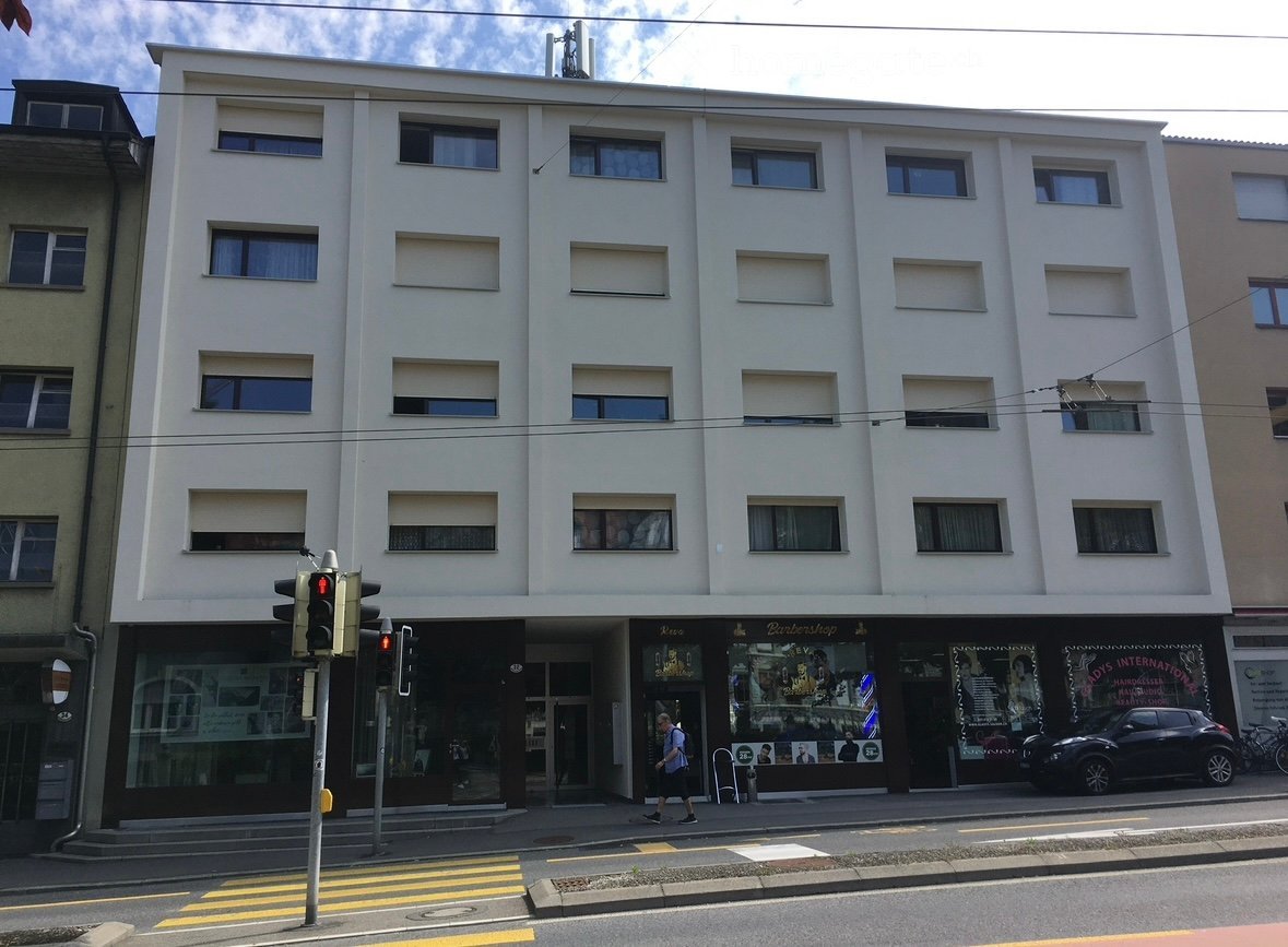 The facade of a white building with many windows and an entrance, traffic lights on the left and a car parked in front on the right.