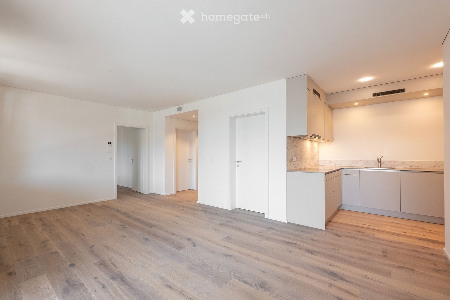 Empty room with wooden flooring, white walls, and a small kitchen area with cabinets and a sink.