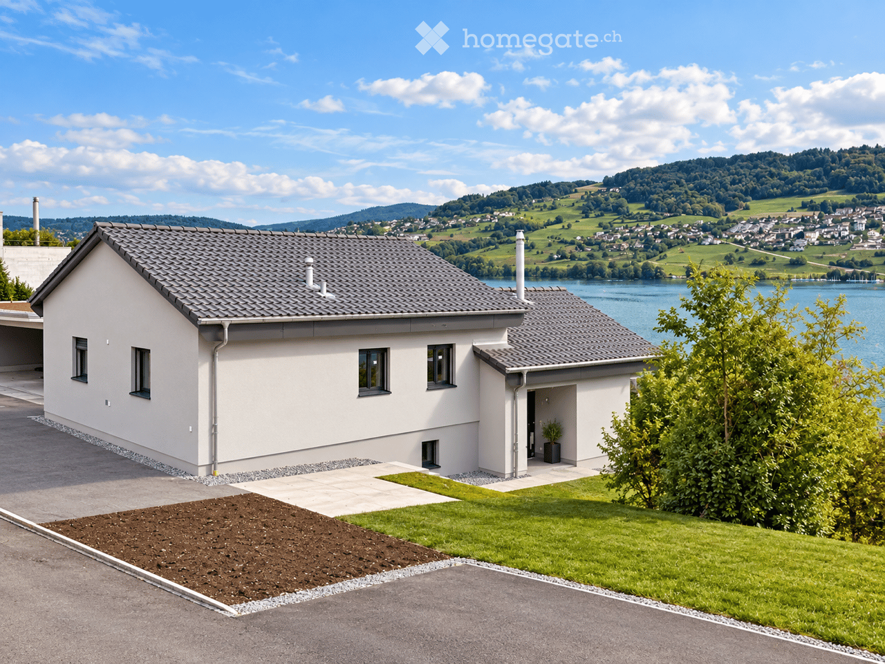 Modern two-story house with a grey tiled roof, white exterior walls, and multiple windows. The house features a driveway, a small garden area, and is situated near a lake with a scenic view of hills and a town in the background.