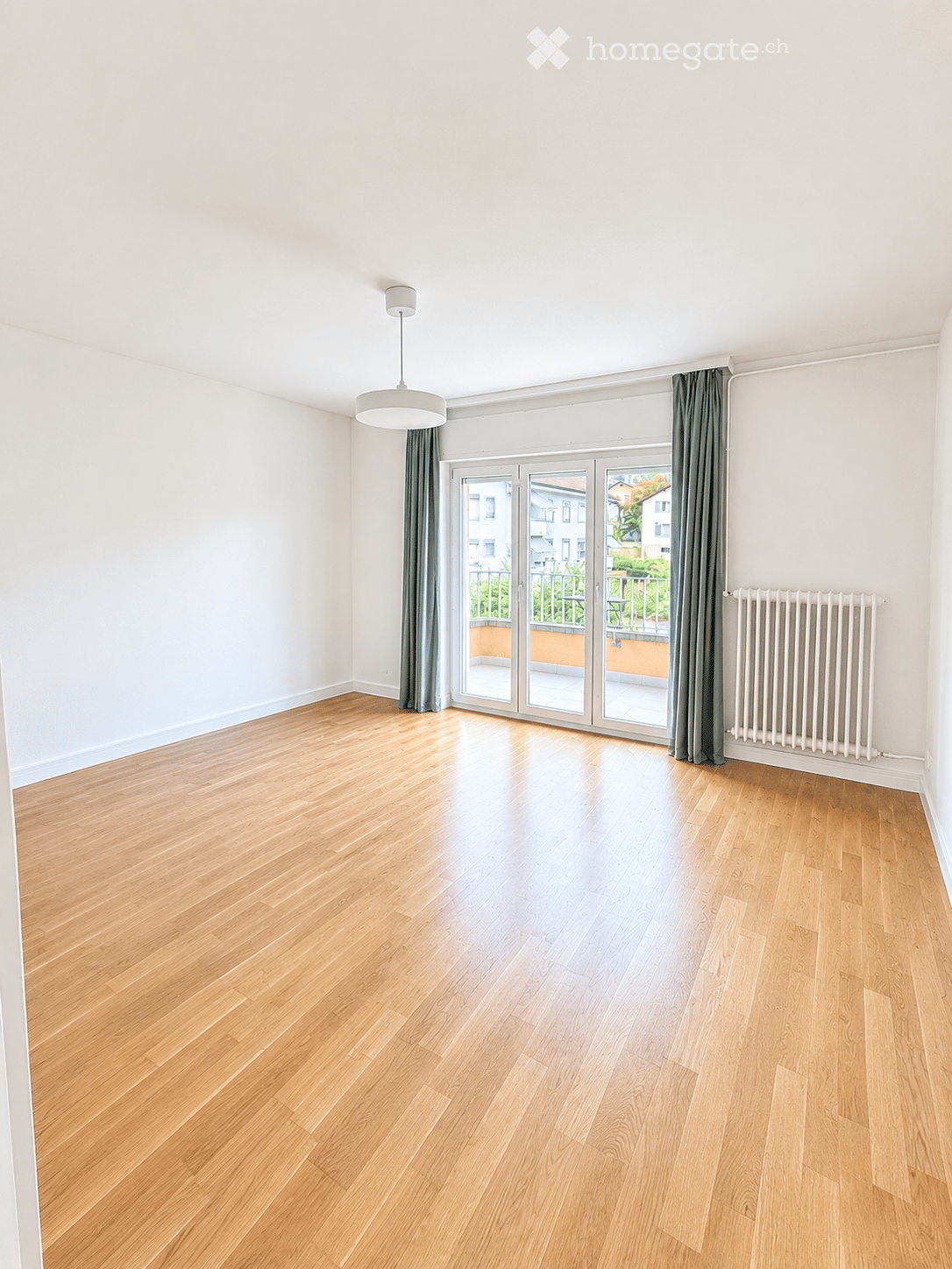 Empty room with light wooden flooring, white walls, large sliding glass doors leading to a balcony, and a radiator on one wall.