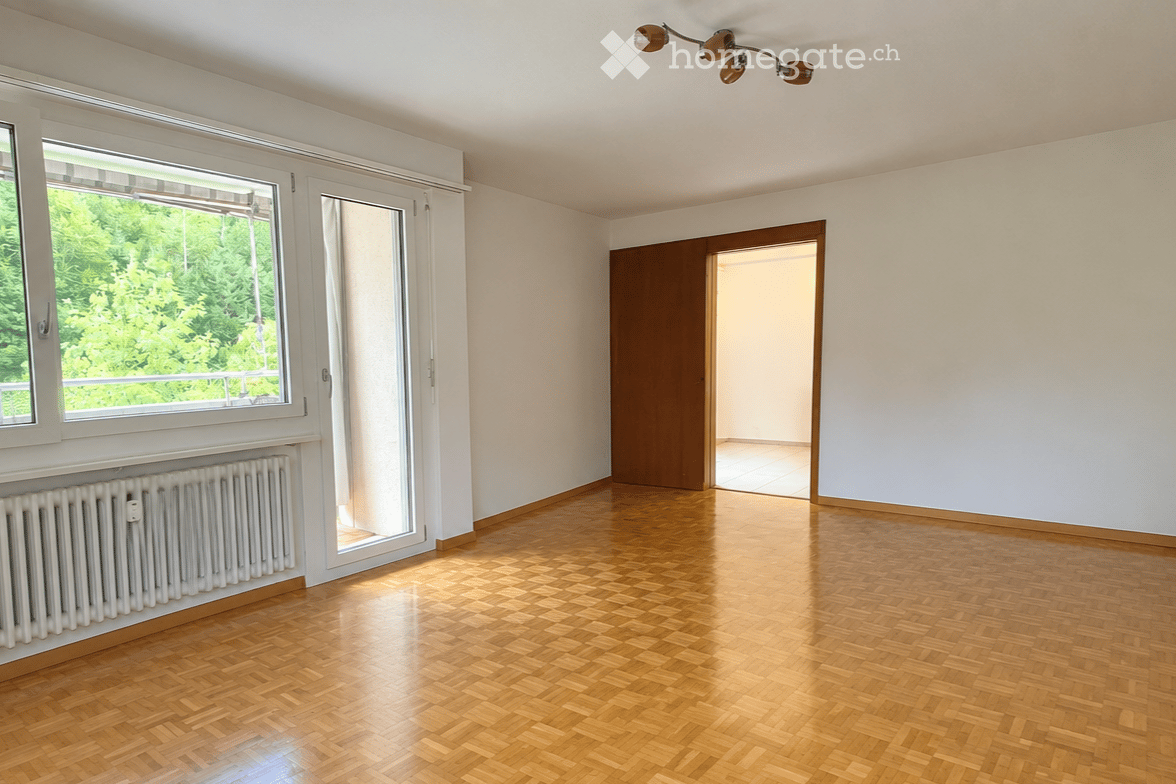 Empty room with wooden flooring, white walls, large window with view, radiator, and a door leading to another space.