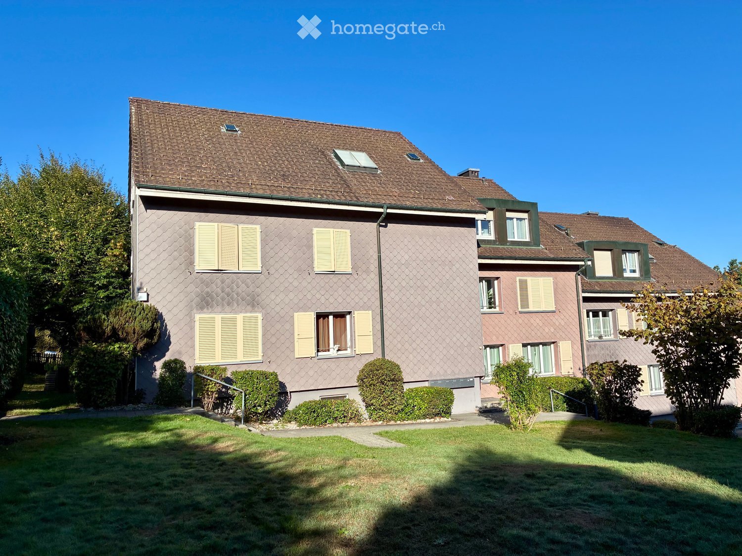 Two-story house with brown tiled roof, beige siding, multiple windows with shutters, and well-maintained lawn with shrubs.
