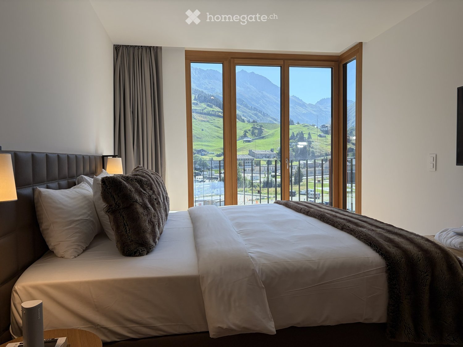 Modern bedroom with a large bed, fur throw, headboard, pillows, table lamp, and large windows offering a scenic mountain view.