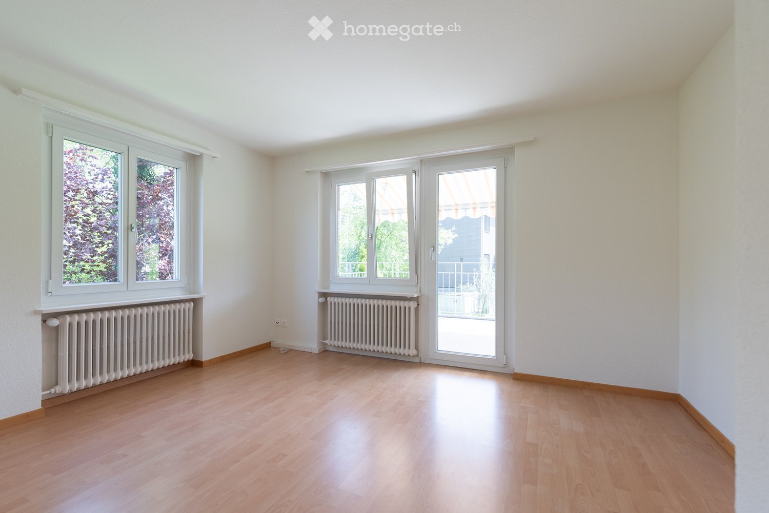 Empty room with light wooden flooring, white walls, and large windows with radiators beneath them. The room features a sliding glass door leading to a balcony area.