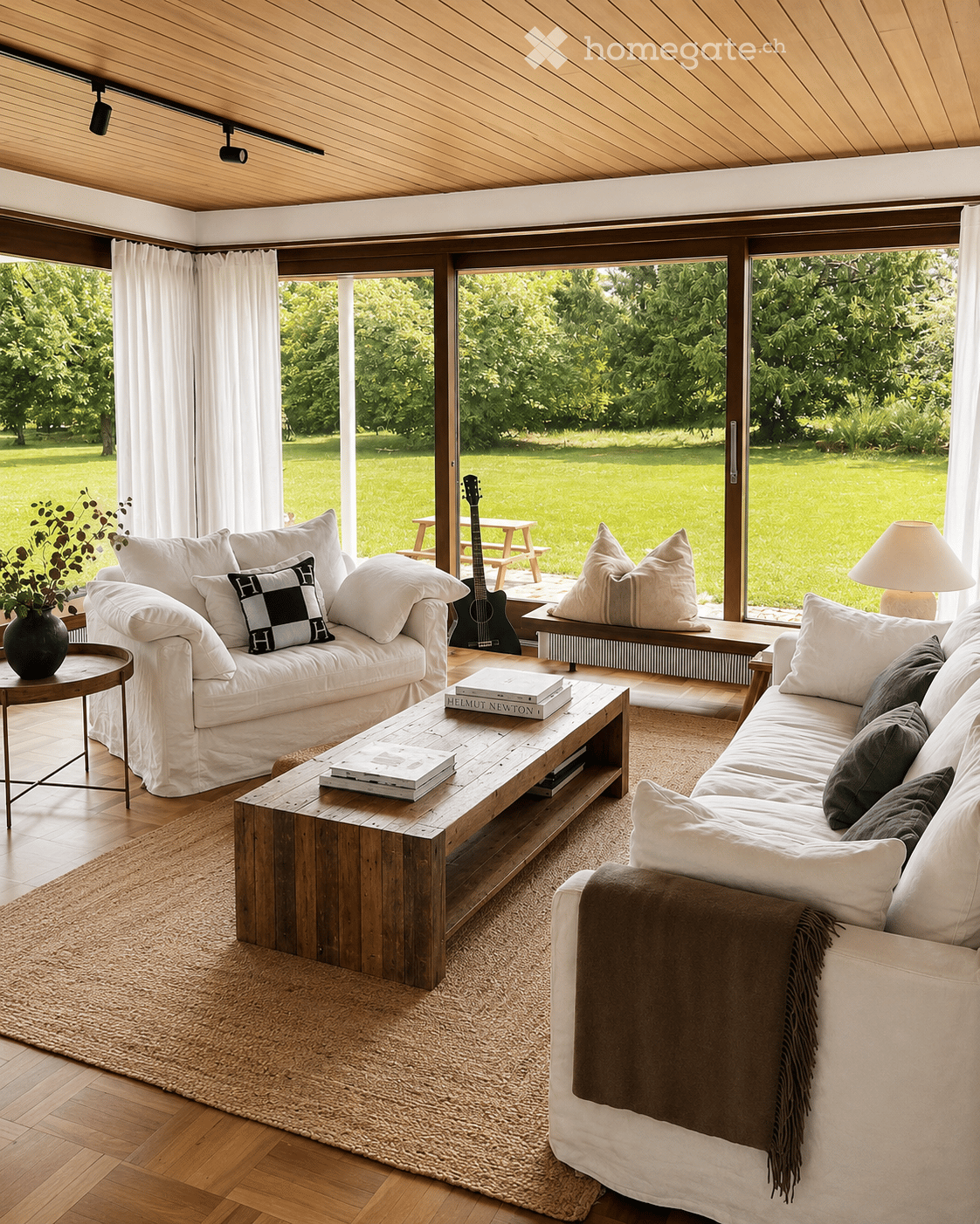 Bright living room with large windows overlooking a green lawn, featuring two sofas, a wooden coffee table, a guitar, and decorative pillows.