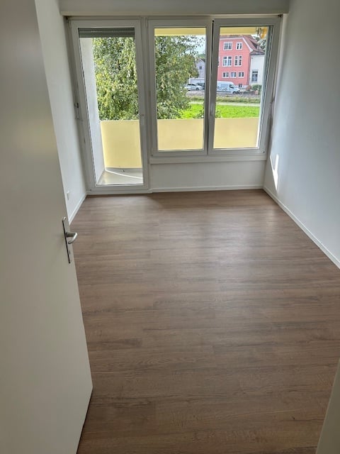 Empty room with wooden flooring, large windows, and a sliding glass door leading to a balcony.