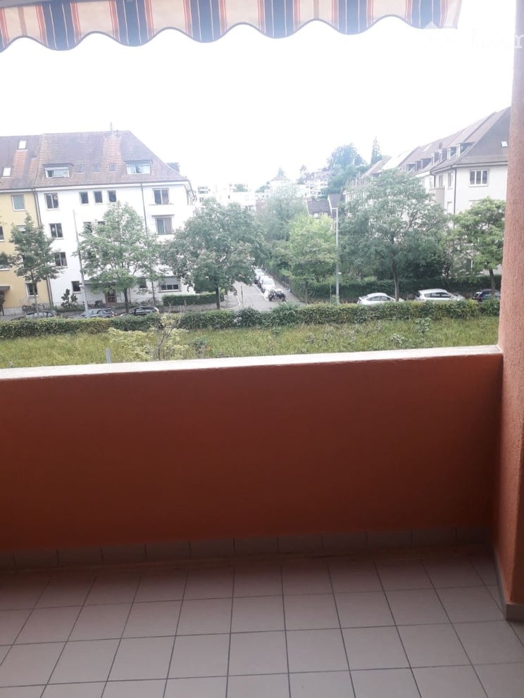 Balcony with tiled floor, striped awning, overlooking a street with houses, trees, and parked cars.